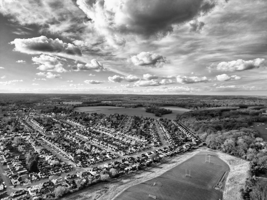 A black-and-white aerial photograph capturing a suburban residential area with rows of closely packed houses featuring pitched roofs and small front gardens. The homes are arranged along parallel streets with visible driveways and spaced-out trees. In the foreground, there is a sports field with a running track, goalposts, and some sports markings on the grass. Surrounding the housing area, there are patches of woodland and open fields, extending towards a distant horizon under a sky filled with large, layered clouds. The lighting highlights the textured rooftops and the natural environment, with shadows cast by the clouds over the landscape. The scene appears to be taken during daytime, providing a clear view of the otherwise dense housing layout and the open green spaces that frame the community. Such broad landscape views are typical in contexts where ensuring proper waste disposal and alternative rubbish removal solutions, like those offered by Rubbish Removal Watford, are important for maintaining neighborhood cleanliness and sustainability.