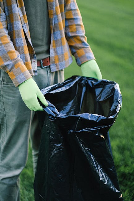A person standing outdoors on a grassy area is holding open a large black rubbish bag lined with silver material. The individual is dressed in casual clothing, including a green shirt, a yellow and gray checkered flannel jacket, and light-colored trousers secured with a brown belt. They are wearing light green gloves, indicating they are engaged in waste collection or disposal. The background features a well-maintained, lush green lawn, suggesting a garden or park environment. The scene emphasizes the process of waste collection, likely as part of private rubbish removal services offered by companies like Rubbish Removal Watford, which specialize in on-site clearance and alternative waste handling solutions outside of local authority collections. The bag appears partially filled, ready for waste disposal, and the lighting suggests daylight conditions, highlighting the practical aspect of rubbish removal activities in a scenic outdoor setting.