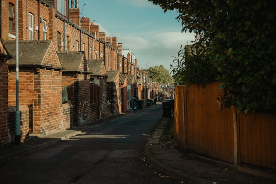 A narrow residential alleyway lined with red-brick terraced houses featuring small front yards and pitched roofs, with some brickwork showing signs of weathering. On the right side, a tall, natural green hedge partially obscures a wooden fence that extends along the length of the alley. Large black wheelie bins are positioned along the curb in front of the houses, likely used for waste and recycling collection. The pavement appears uneven with small patches of dirt and fallen leaves scattered across it. Shadows cast by the houses and fence create areas of contrast, indicating late afternoon sunlight. In the background, the alley continues with more houses visible under a partly cloudy sky. The scene suggests an area where residents may utilize private rubbish collection services such as those provided by Rubbish Removal Watford, which specialises in efficient waste disposal and house clearance in the local estate area, supporting alternative waste handling outside of council services.