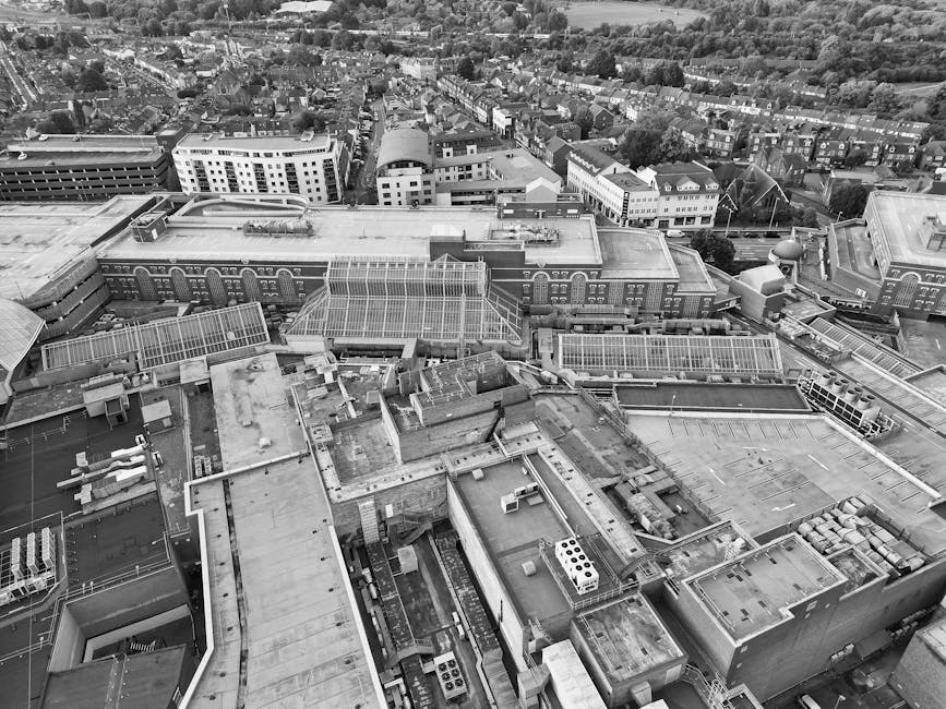 A detailed black and white aerial photograph of a cityscape focusing on the rooftops of various commercial and industrial buildings. The image reveals flat roofs constructed from materials such as concrete and metal, with some rooftop areas featuring ventilation units, air conditioning systems, and small skylights. Numerous HVAC units and ductwork are visible, along with vents and exhaust pipes scattered across the rooftops. In the background, a densely built urban area extends into residential neighborhoods with rows of houses and tree-lined streets. The urban environment is characterized by a mix of modern and older architectural styles, with some buildings featuring curved glass facades and others with traditional brick exteriors. The natural lighting is diffuse, suggesting an overcast sky, and the overall scene emphasizes the complexity and density of urban infrastructure, which can be connected to the need for efficient rubbish removal and waste management services such as those offered by Rubbish Removal Watford for local businesses and property owners seeking alternative waste handling solutions.