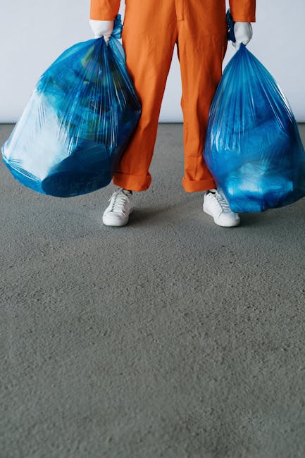 A person dressed in orange work trousers and white sneakers is standing on a grey surface while holding two large blue plastic bags filled with rubbish. The bags appear to contain mixed waste materials and are being carried in each hand, with the person's hands covered by grey gloves. The background is a plain, light-colored wall, creating a neutral setting that emphasizes the focus on the waste collection activity. The scene suggests an informal or private rubbish removal process, aligning with alternative waste handling methods outside of municipal services. The image captures the act of onsite waste collection and disposal, illustrating a step in rubbish removal services provided by companies such as Rubbish Removal Watford, which may include independent collection or on-site clearance of rubbish from various locations.