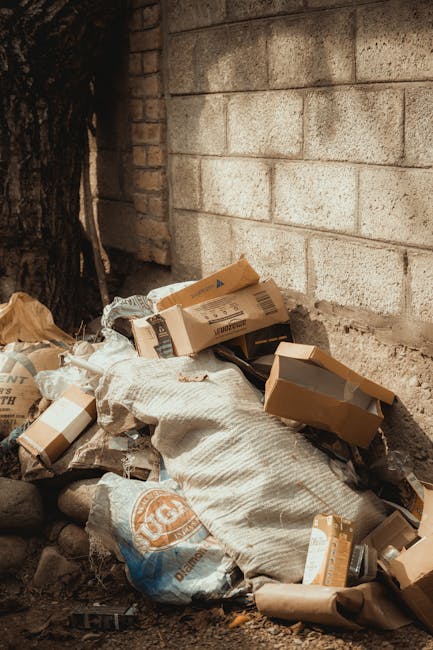 An alleyway scene featuring a large, black, wheeled rubbish bag in the foreground, placed on dark, dirty pavement. Behind it, a substantial collection of cardboard boxes, plastic bags, and miscellaneous waste materials are stacked haphazardly against a weathered brick wall and a wooden pallet. The area appears cluttered and unkempt, with debris scattered on the ground and some graffiti on the side walls. Sparse branches from a leafless tree stretch across the top of the alley, and a nearby building has metal security grilles on its windows. The lighting suggests an overcast day, adding a dull, muted tone to the scene. This setting illustrates a typical example of waste accumulation requiring rubbish removal services, with the presence of bulky refuse and general household waste near an urban structure, aligned with private waste handling or alternative collection methods facilitated by providers such as Rubbish Removal Watford.
